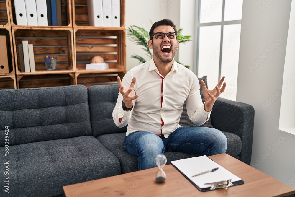 Young hispanic man with beard working at consultation office crazy and mad shouting and yelling with aggressive expression and arms raised. frustration concept. Young hispanic man with beard working at consultation office crazy and mad shouting and yelling with aggressive expression and arms raised. frustration concept.