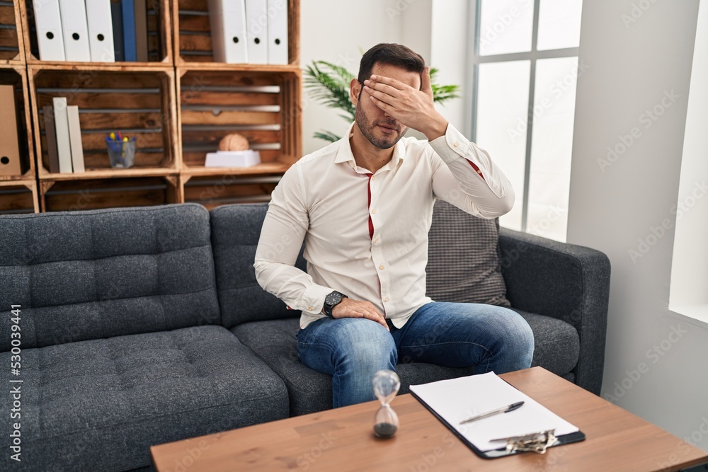 Young hispanic man with beard working at consultation office covering eyes with hand, looking serious and sad. sightless, hiding and rejection concept Young hispanic man with beard working at consultation office covering eyes with hand, looking serious and sad. sightless, hiding and rejection concept