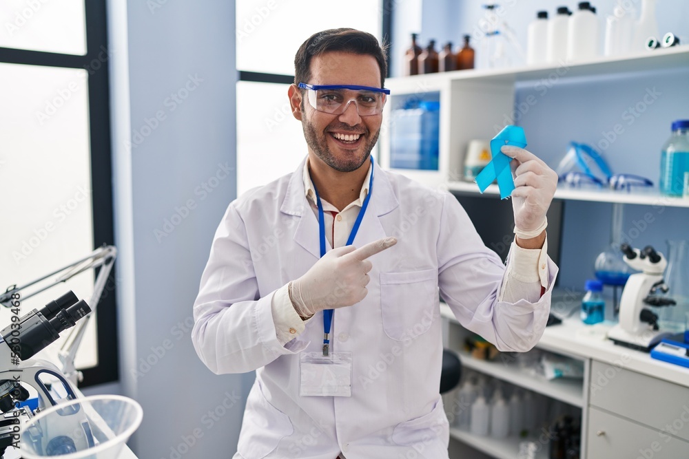 Young hispanic man with beard working at scientist laboratory holding blue ribbon smiling happy pointing with hand and finger Young hispanic man with beard working at scientist laboratory holding blue ribbon smiling happy pointing with hand and finger