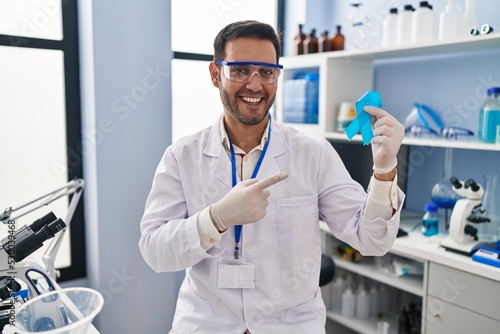 Young hispanic man with beard working at scientist laboratory holding blue ribbon smiling happy pointing with hand and finger