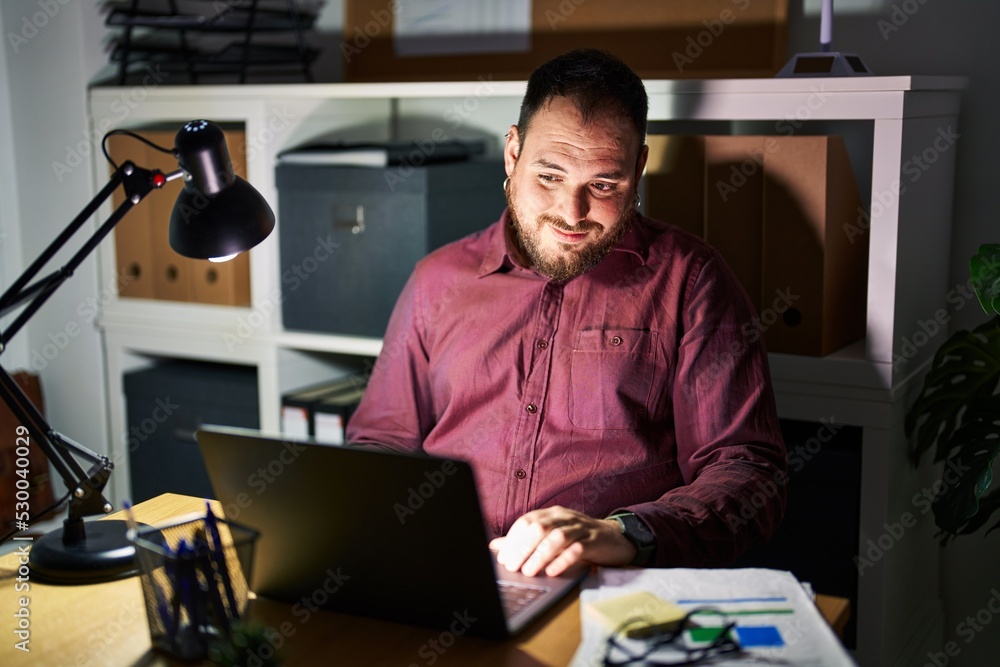 Plus size hispanic man with beard working at the office at night looking away to side with smile on face, natural expression. laughing confident. Plus size hispanic man with beard working at the office at night looking away to side with smile on face, natural expression. laughing confident.