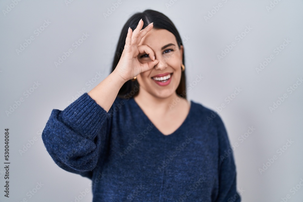 Young brunette woman standing over isolated background doing ok gesture with hand smiling, eye looking through fingers with happy face. Young brunette woman standing over isolated background doing ok gesture with hand smiling, eye looking through fingers with happy face.