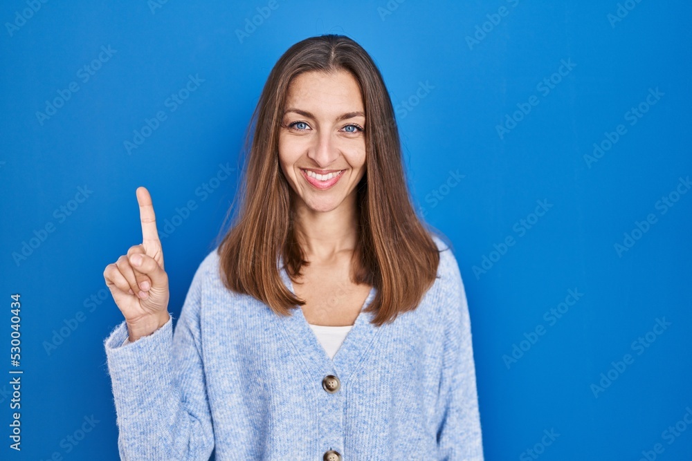 Young woman standing over blue background showing and pointing up with finger number one while smiling confident and happy. Young woman standing over blue background showing and pointing up with finger number one while smiling confident and happy.