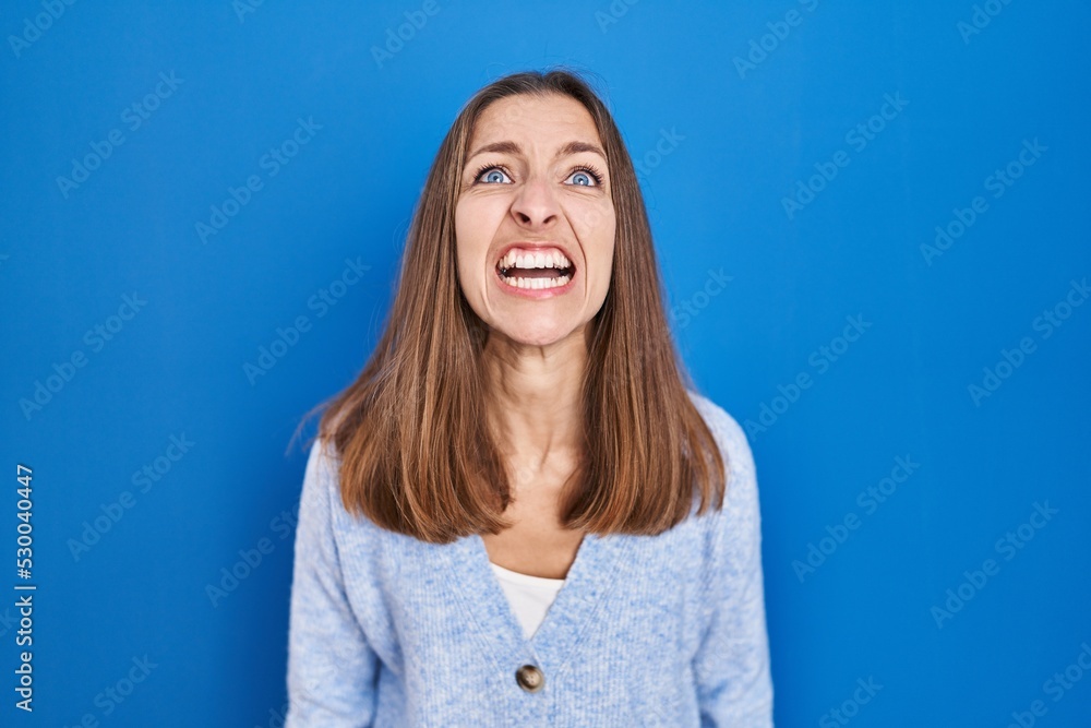 Young woman standing over blue background angry and mad screaming frustrated and furious, shouting with anger. rage and aggressive concept. Young woman standing over blue background angry and mad screaming frustrated and furious, shouting with anger. rage and aggressive concept.