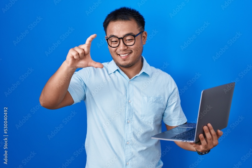Chinese young man using computer laptop smiling and confident gesturing with hand doing small size sign with fingers looking and the camera. measure concept. Chinese young man using computer laptop smiling and confident gesturing with hand doing small size sign with fingers looking and the camera. measure concept.