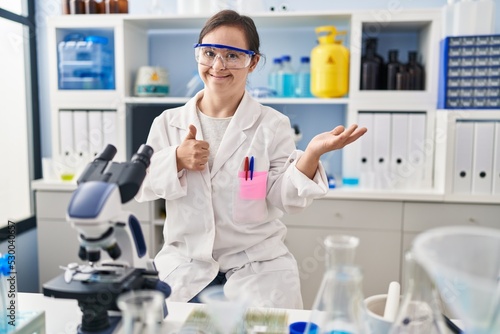 Hispanic girl with down syndrome working at scientist laboratory showing palm hand and doing ok gesture with thumbs up  smiling happy and cheerful