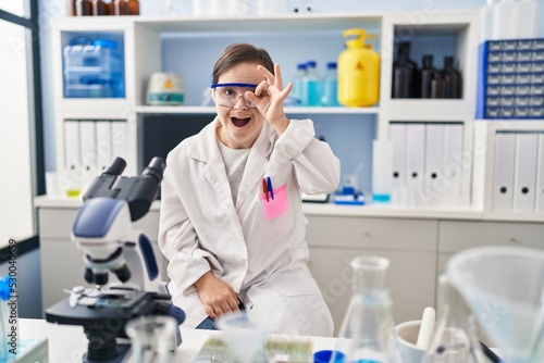 Hispanic girl with down syndrome working at scientist laboratory doing ok gesture shocked with surprised face  eye looking through fingers. unbelieving expression.
