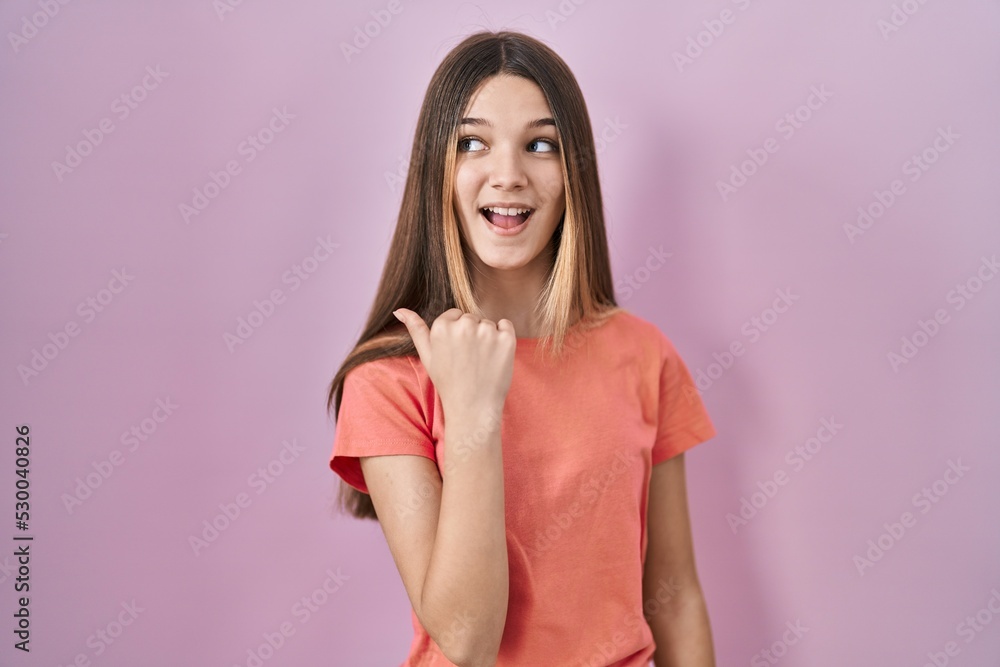 Teenager girl standing over pink background smiling with happy face looking and pointing to the side with thumb up. Teenager girl standing over pink background smiling with happy face looking and pointing to the side with thumb up.