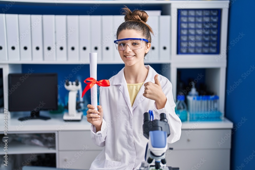 Teenager girl working at scientist laboratory holding degree smiling happy and positive, thumb up doing excellent and approval sign Teenager girl working at scientist laboratory holding degree smiling happy and positive, thumb up doing excellent and approval sign