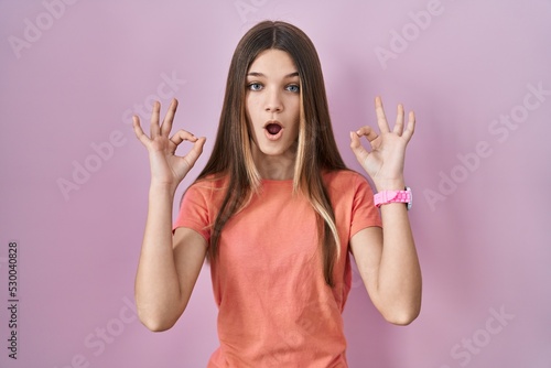 Teenager girl standing over pink background looking surprised and shocked doing ok approval symbol with fingers. crazy expression