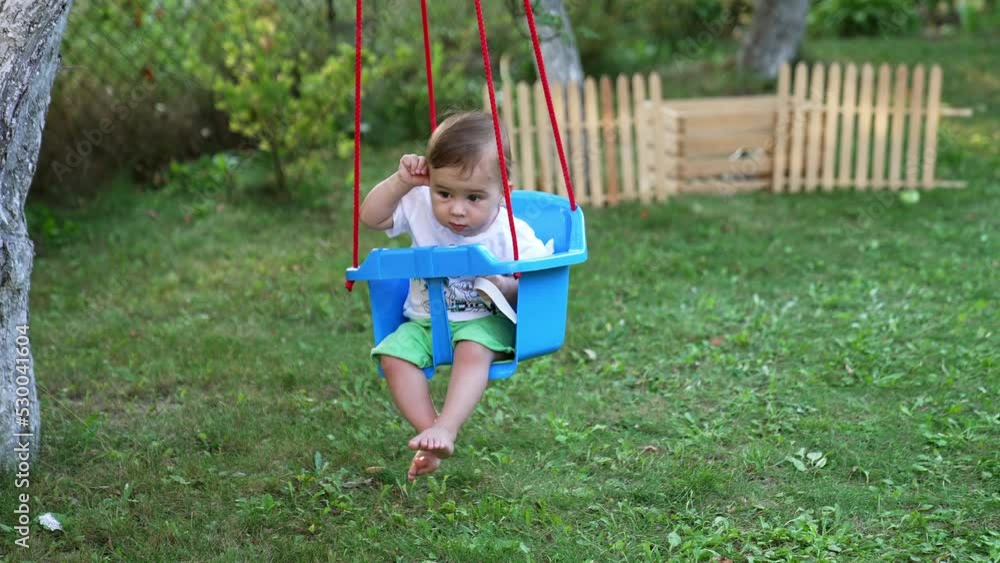 Little kid in t-shirt and shorts swaying in a swing. Calm barefoot baby boy in the garden in summer. Nature backdrop in blur. Little kid in t-shirt and shorts swaying in a swing. Calm barefoot baby boy in the garden in summer. Nature backdrop in blur.