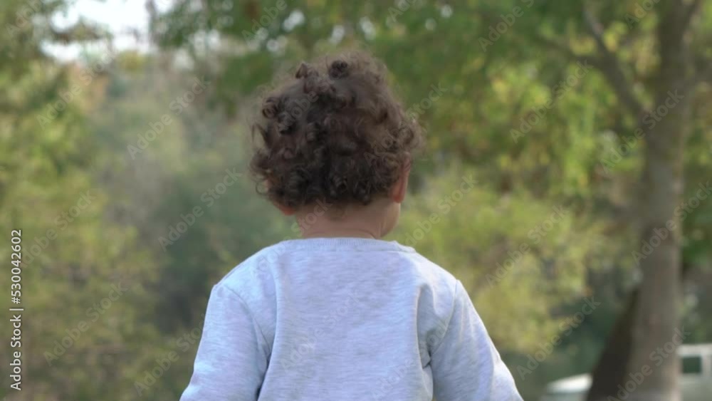 Little child with cute curly hair, walks away in grassy sycamore forest. There are a white station wagon and woman in the background. Little child with cute curly hair, walks away in grassy sycamore forest. There are a white station wagon and woman in the background.