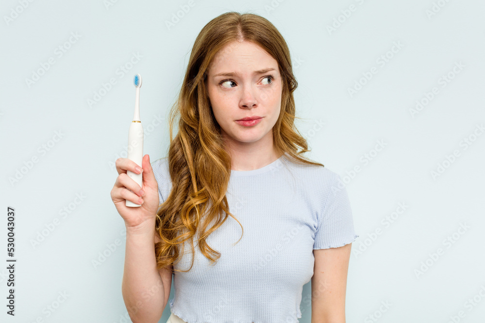 Young caucasian woman holding electric toothbrush isolated on blue background confused, feels doubtful and unsure. Young caucasian woman holding electric toothbrush isolated on blue background confused, feels doubtful and unsure.