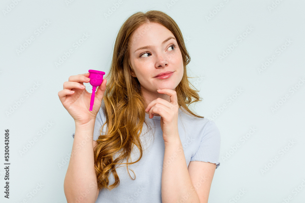 Young caucasian woman holding menstrual cup isolated on blue background looking sideways with doubtful and skeptical expression. Young caucasian woman holding menstrual cup isolated on blue background looking sideways with doubtful and skeptical expression.