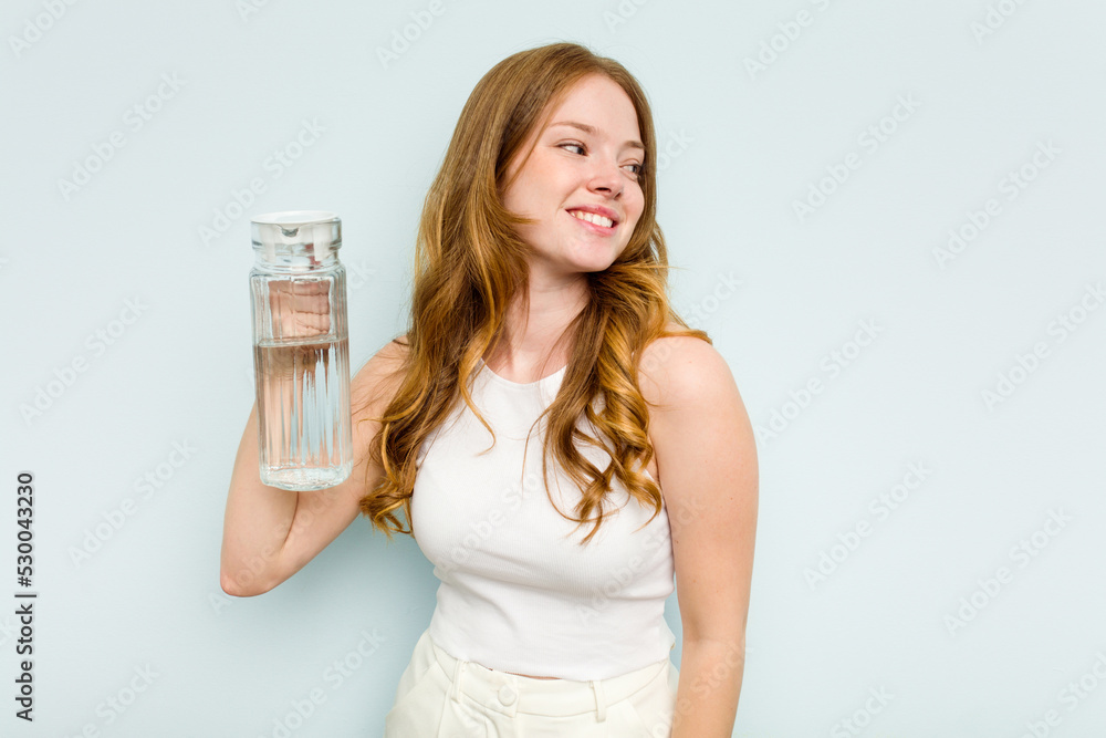 Young caucasian woman holding jar of water isolated on blue background looks aside smiling, cheerful and pleasant. Young caucasian woman holding jar of water isolated on blue background looks aside smiling, cheerful and pleasant.