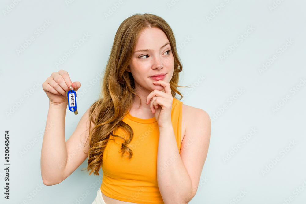 Young caucasian woman holding home keys isolated on blue background relaxed thinking about something looking at a copy space. Young caucasian woman holding home keys isolated on blue background relaxed thinking about something looking at a copy space.