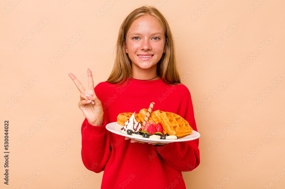 Little caucasian girl holding a waffles isolated on beige background showing number two with fingers. Little caucasian girl holding a waffles isolated on beige background showing number two with fingers.