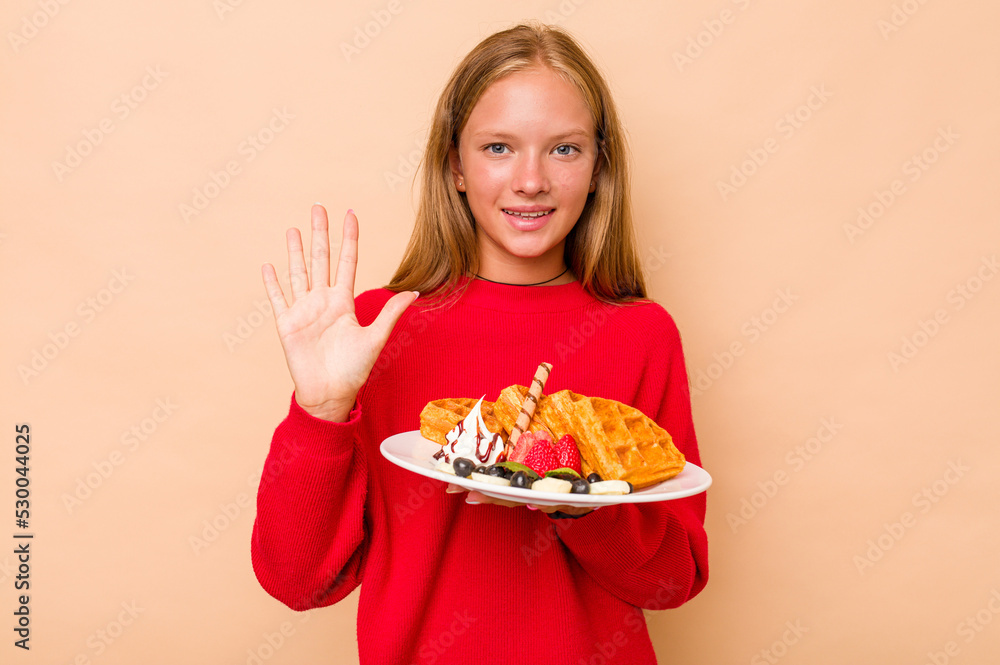 Little caucasian girl holding a waffles isolated on beige background smiling cheerful showing number five with fingers. Little caucasian girl holding a waffles isolated on beige background smiling cheerful showing number five with fingers.