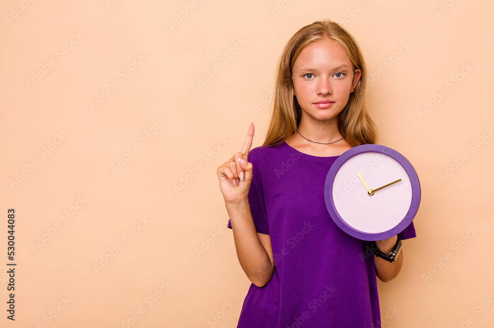 Little caucasian girl holding a clock isolated on beige background showing number one with finger. Little caucasian girl holding a clock isolated on beige background showing number one with finger.