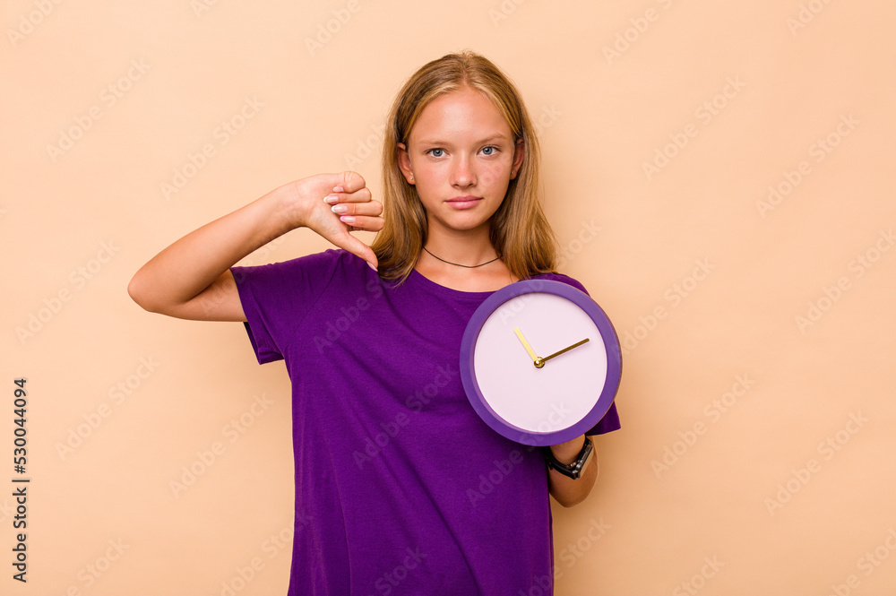Little caucasian girl holding a clock isolated on beige background showing a dislike gesture, thumbs down. Disagreement concept. Little caucasian girl holding a clock isolated on beige background showing a dislike gesture, thumbs down. Disagreement concept.