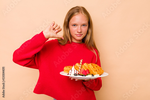 Little caucasian girl holding a waffles isolated on beige background feels proud and self confident, example to follow.