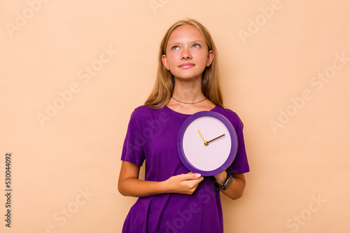 Little caucasian girl holding a clock isolated on beige background dreaming of achieving goals and purposes