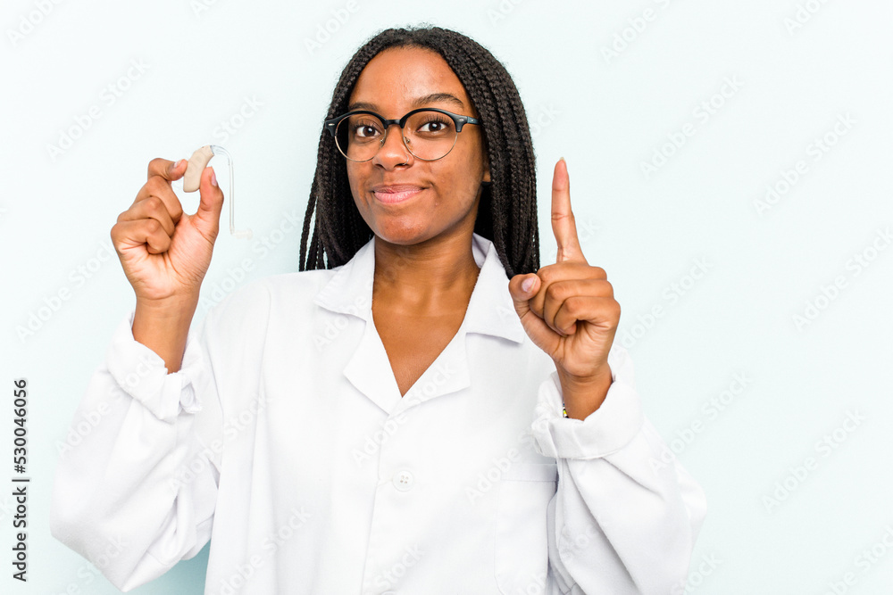 Young African American otorhinolaryngologist woman holding a hearing aid isolated on blue background showing number one with finger. Young African American otorhinolaryngologist woman holding a hearing aid isolated on blue background showing number one with finger.