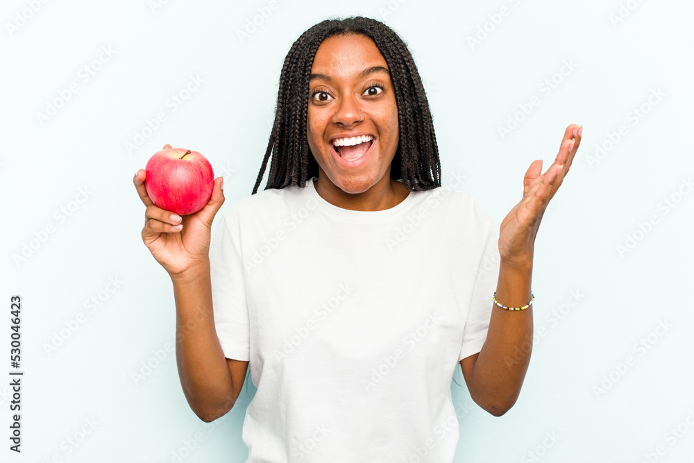 Young African American woman holding an apple isolated on blue background receiving a pleasant surprise, excited and raising hands.