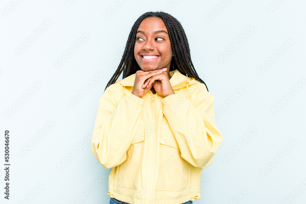 Young African American woman with braids hair isolated on blue background keeps hands under chin, is looking happily aside. Young African American woman with braids hair isolated on blue background keeps hands under chin, is looking happily aside.