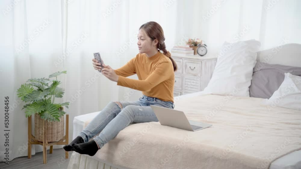 Asian woman sitting on bed taking selfie while using laptop working. Happy female making video call relatives or bestfriend with smartphone at home. Concept of technology.
Asian woman sitting on bed taking selfie while using laptop working. Happy female making video call relatives or bestfriend with smartphone at home. Concept of technology.