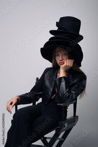 brunette model in leather jacket and different black hats sitting on chair isolated on grey.