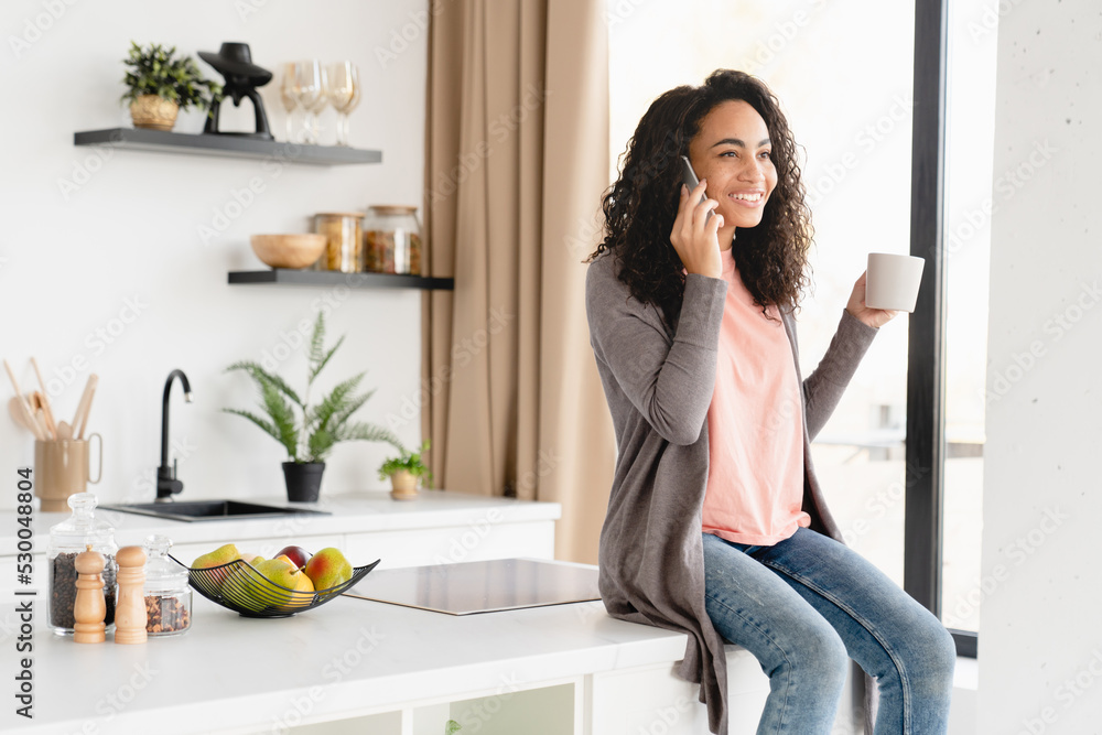 African young woman housewife freelancer having breakfast drinking coffee tea at home kitchen while talking on smart phone with friends on mobile application