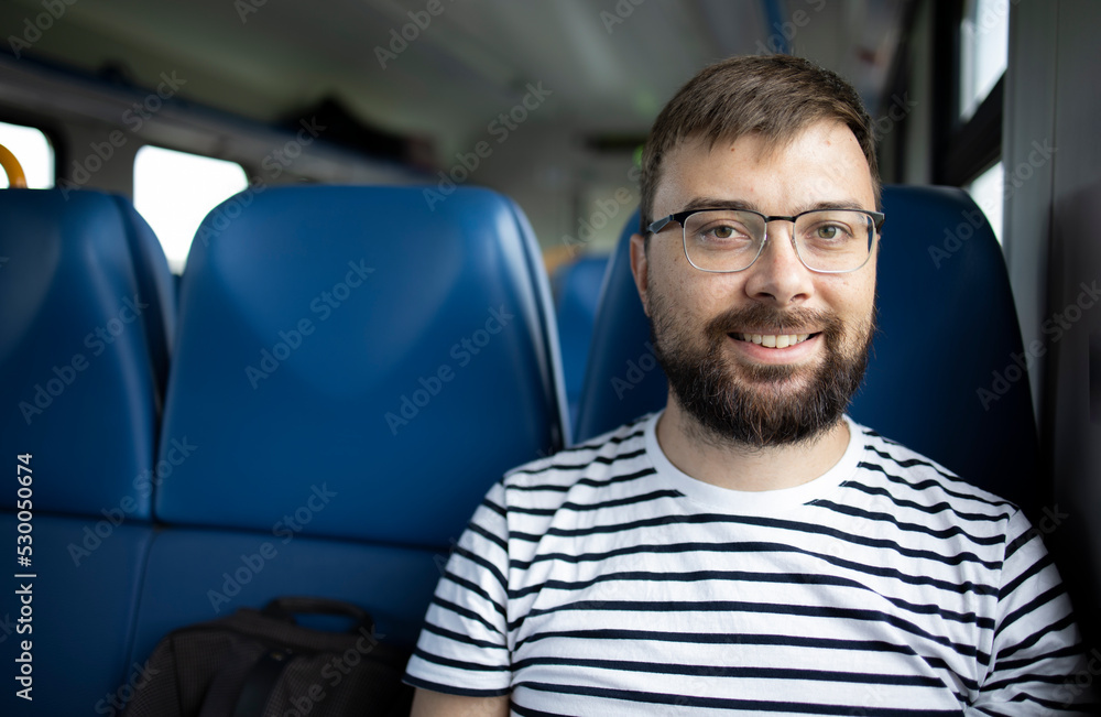 Handsome tourist guy in glasses and a striped t-shirt with a backpack looks directly at the camera in an electric train car. Travel by train. Concept: travel, weekend, vacation Handsome tourist guy in glasses and a striped t-shirt with a backpack looks directly at the camera in an electric train car. Travel by train. Concept: travel, weekend, vacation