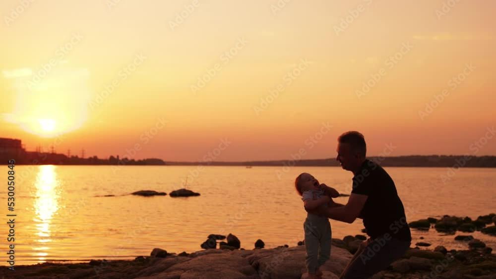 Father entertaining his little toddler boy near the river. Dad is swaying and tossing his son and child is happy. Sunset backdrop. Father entertaining his little toddler boy near the river. Dad is swaying and tossing his son and child is happy. Sunset backdrop.
