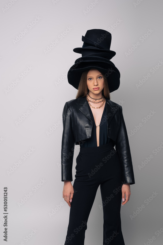 teenage model in leather jacket and different black hats on head isolated on grey. teenage model in leather jacket and different black hats on head isolated on grey.