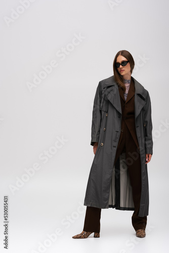 full length of stylish teenage model in sunglasses and trendy trench coat posing on grey.