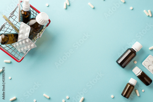 Illness concept. Top view photo of shopping cart with medicines transparent brown bottles pills blisters capsules and thermometer on isolated pastel blue background with copyspace