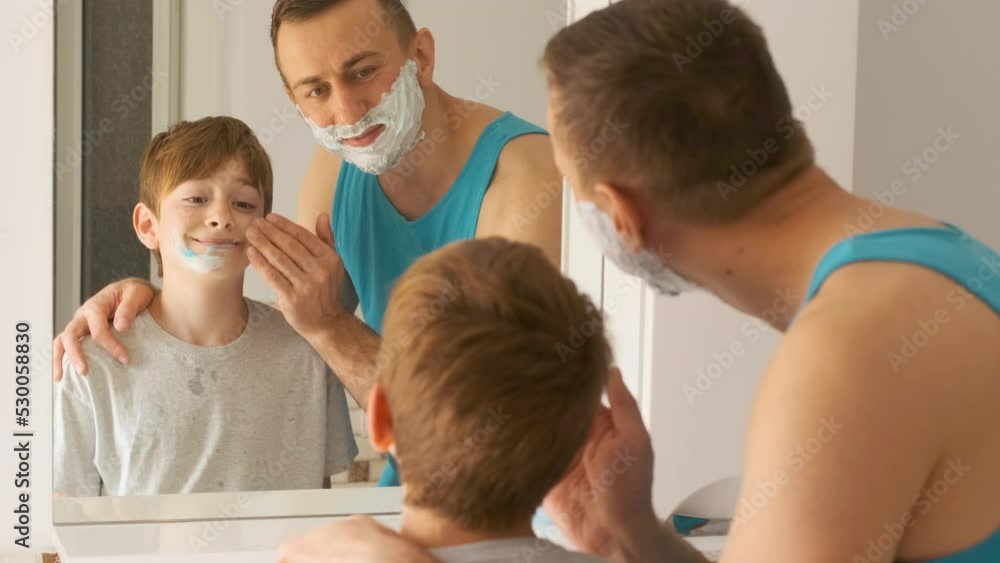 Father with schoolboy son looks into reflection in mirror and man applies shaving foam to boys face Father with schoolboy son looks into reflection in mirror and man applies shaving foam to boys face