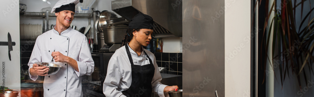 cheerful chef whisking eggs and looking at african american colleague in kitchen, banner. cheerful chef whisking eggs and looking at african american colleague in kitchen, banner.