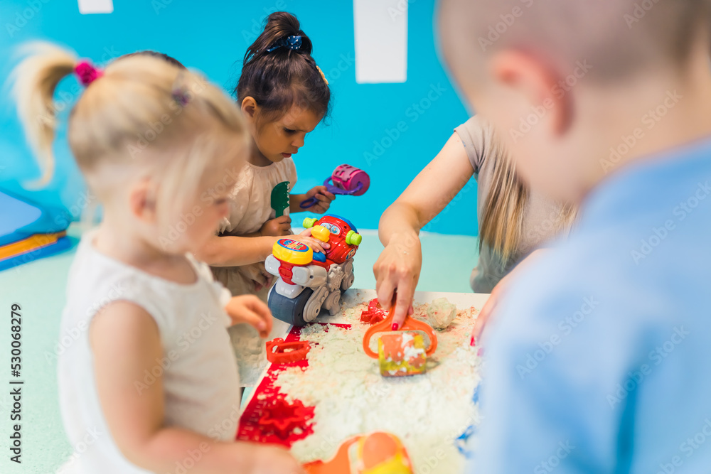 Relaxing sensory play with moldable kinetic sand at nursery school. Toddlers with their teacher having fun around the table using different tools for sculpting sand such as colorful and textured - obrazy, fototapety, plakaty 