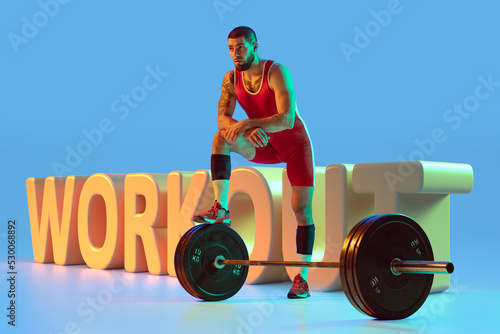 Workout. Young muscular athlete posing with 10kg barbell over huge 3d lettering on blue background in neon light. Sport, weightlifting, power, strength photo