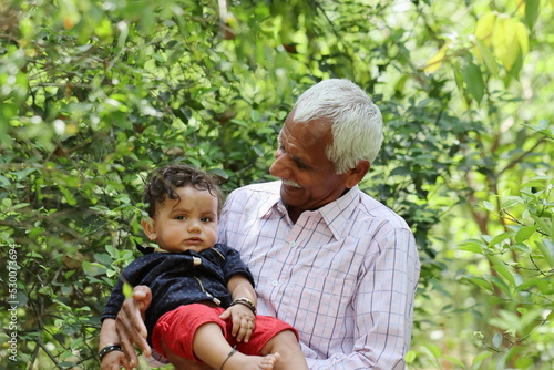 Picture of a proud grandfather holding his Adorable grandson and looking at him with love. Standing in a backyard of green a farm.