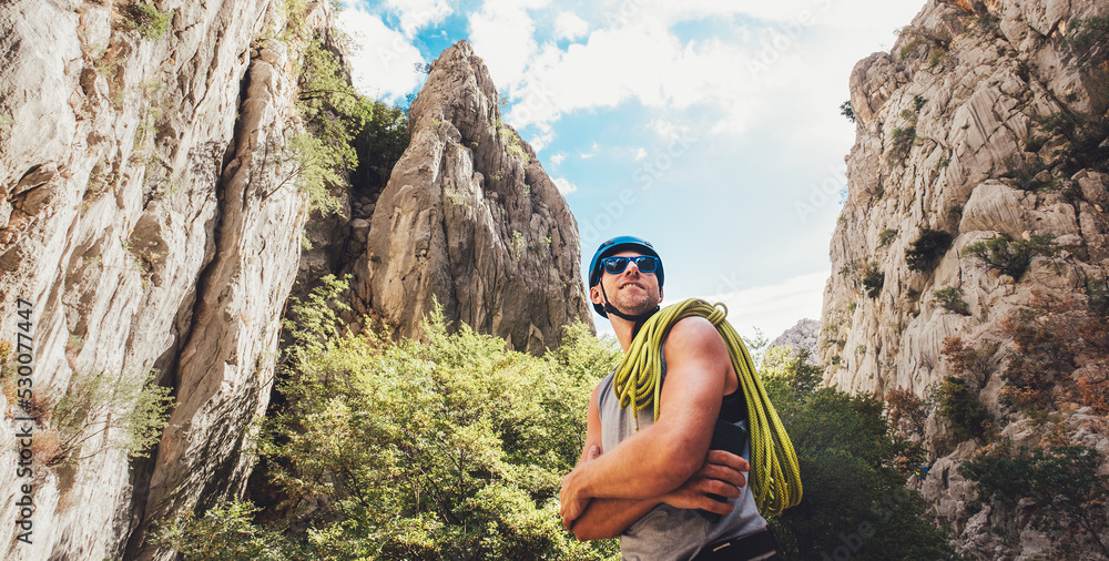 Portrait of smiling climber man in protective helmet and sunglasses with climbing rope on the shoulder standing in Paklenica park between rock cliff walls. Active extreme sports time spending concept.