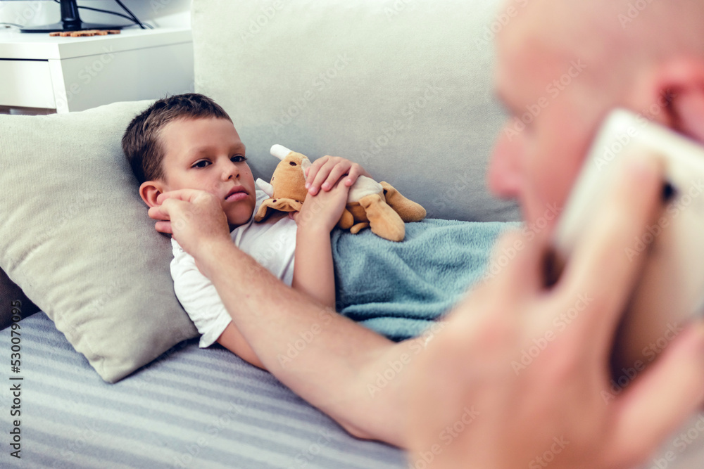 Worried man talking on smartphone while sitting near diseased son in the dedroom. Worried father consulting doctor on cellphone while sitting next to his little son in the living room. Worried man talking on smartphone while sitting near diseased son in the dedroom. Worried father consulting doctor on cellphone while sitting next to his little son in the living room.