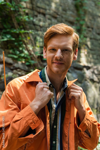 joyful redhead man in orange jacket smiling and adjusting collar on jacket outside.