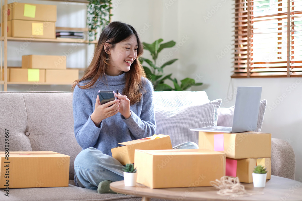 Young beautiful happy asian business woman owner of SME online using laptop and smartphone receive order from customer with parcel box packaging at her startup home office, online business seller Young beautiful happy asian business woman owner of SME online using laptop and smartphone receive order from customer with parcel box packaging at her startup home office, online business seller
