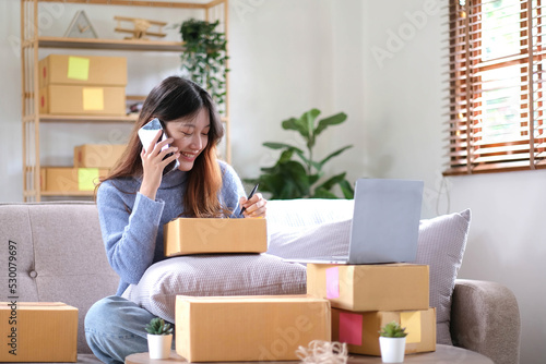 Portrait young asian woman using laptop computer for work with parcel box on sofa in living room interior