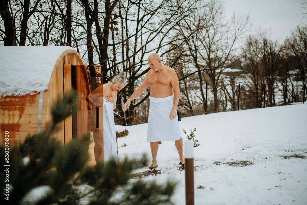 Senior woman in towel with her husband coming out from outdoor sauna during cold winter day. - obrazy, fototapety, plakaty 
