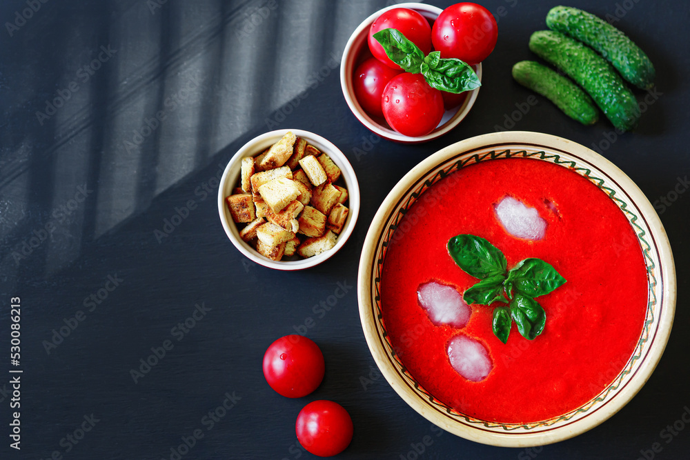 Red Spanish vegetable cold cream soup gazpacho in plate with basil leaves, ice cubes in it, ingredients around: tomatoes, cucumbers, croutons; shadow from blinds, flatlay, horizontal, dark background Red Spanish vegetable cold cream soup gazpacho in plate with basil leaves, ice cubes in it, ingredients around: tomatoes, cucumbers, croutons; shadow from blinds, flatlay, horizontal, dark background