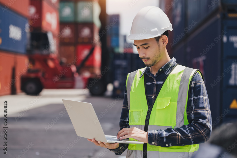 Portrait smart Asian engineer male wearing safety vest and hard helmet hand holding laptop computer standing in front of container box on site Cargo freight ship for import and export. Transport man Portrait smart Asian engineer male wearing safety vest and hard helmet hand holding laptop computer standing in front of container box on site Cargo freight ship for import and export. Transport man